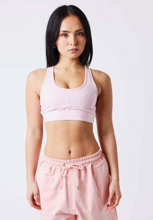 Femme aux cheveux noirs lisses portant un soutien-gorge de sport rose clair et un pantalon de survêtement assorti à cordon, debout devant un fond clair uni.