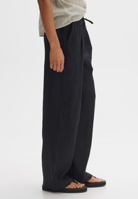 Weite schwarze Hosen aus leichtem Stoff, mit einem elastischen Bund und einem Kordelzug, kombiniert mit flachen Sandalen.