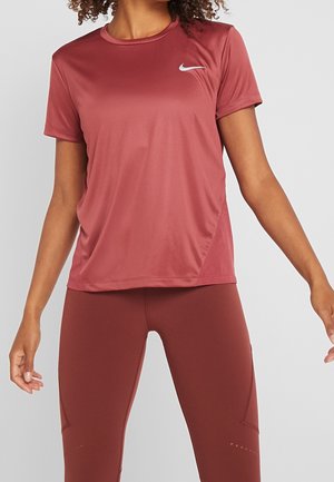 Kobieta ubrana w bordowy krótki rękaw Nike dri-fit i pasujące legginsy, stojąca z lekko rozszerzonymi ramionami.