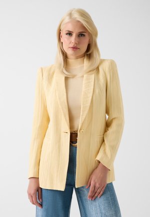Vrouw met blond haar met een lichtgeel gestructureerd blazer over een bijpassende top en een blauwe spijkerbroek, staand voor een effen lichte achtergrond.