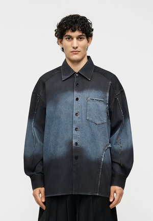 Νέος άνδρας με σγουρά μαλλιά που φοράει ένα πουκάμισο κουμπωτό με μαύρο και μπλε διαβαθμισμένο χρώμα, με ορατές λεπτομέρειες ραφής και τσέπη στο στήθος.