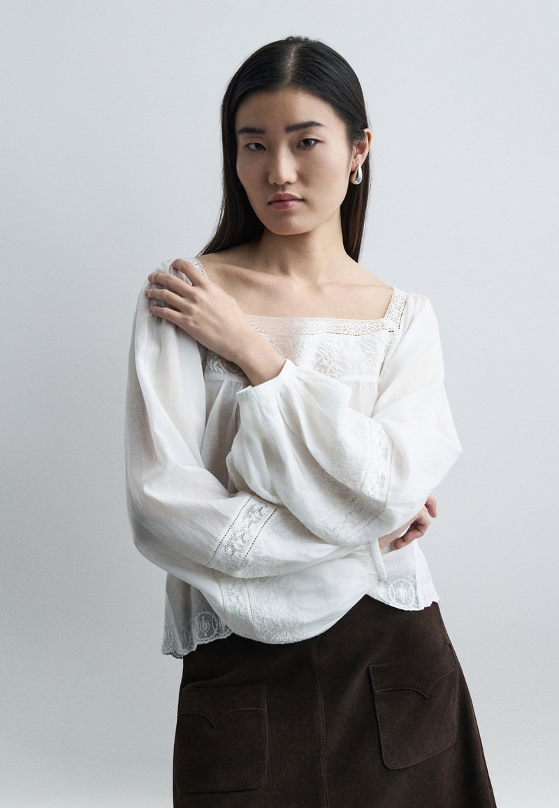 Jeune femme aux longs cheveux noirs portant un chemisier blanc brodé à manches volumineuses et une jupe sombre, posant avec un bras croisé.