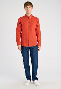 Czerwona koszula na guziki z długimi rękawami, teksturowana tkanina, kieszeń na piersi, zestawiona z ciemnoniebieskimi jeansami i białymi tenisówkami, stojąca na jednolitym tle.