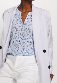 Helles graues Blazer mit schwarzen Knöpfen, getragen über einer blauen Bluse mit Blumenmuster in rosa und dunkelblauen Akzenten; kombiniert mit weißen Hosen.