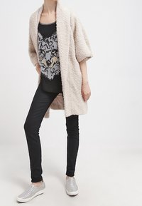 Ljusbeige, fluffig lång kappa över en svart tanktop med en vargs grafik. Svarta slim-fit jeans och silverfärgade slip-on skor.