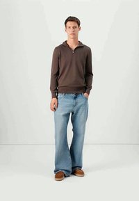 Junger Mann steht da und trägt einen braunen Pullover mit Viertelreißverschluss, hellblaue weite Jeans und braune Slippers vor einem schlichten Hintergrund.