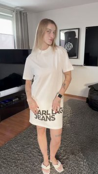 Young person with long blonde hair wearing a white oversized T-shirt dress with "KARL LAGER JEANS" text, standing in a living room.