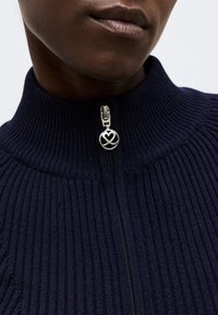 Κοντινό πλάνο ενός ατόμου που φοράει μπλε σκούπο ριμπ πλεκτό πουλόβερ με ασημένιο φερμουάρ διακοσμημένο με καρδιά και κυκλικό σχέδιο.