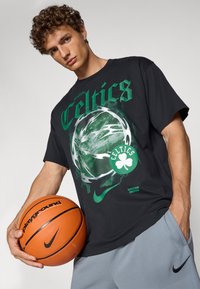 Sort bomulds t-shirt med et grønt Boston Celtics grafik, der inkluderer en kløver og stiliseret tekst. Modellen holder en orange Nike basketball.