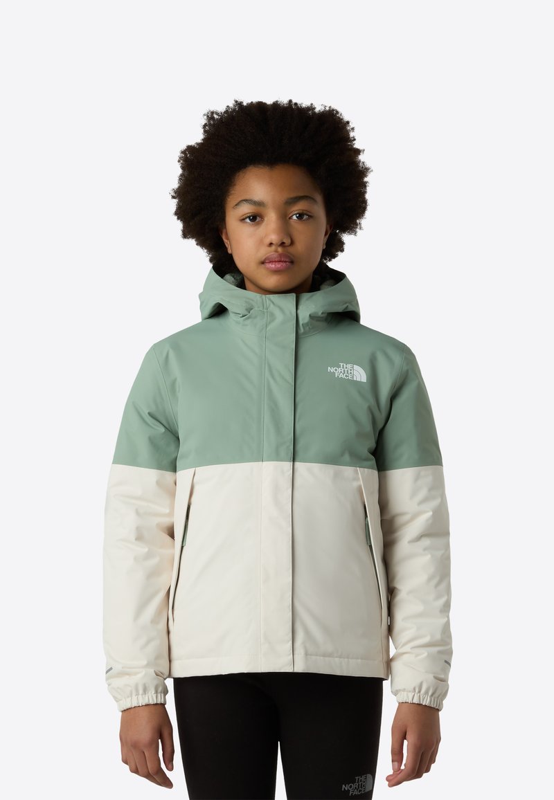 Jeune personne portant une veste d'hiver The North Face verte et blanche avec une capuche, debout devant un fond uni.