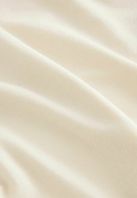 Kermanvärinen kangas, jolla on sileä, silkinen rakenne, esitellen hienovaraisia poimuja ja lempeitä taitteita, korostaen kevyttä, valuvaa laatua.