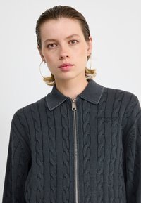 Grijze zip-up trui met kabelbreisel, een kraag en een prominente logo op de borst. Glad textuur met een aansluitende pasvorm.