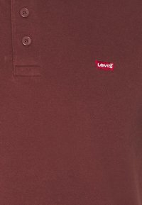 Camisa polo burdeos hecha de tejido texturizado con tres botones marrones; presenta un logo rojo de Levi's en el pecho.