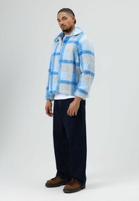 Fleece jacket in light blue and beige check pattern, featuring a collar, button closure, and paired with dark denim pants and brown boots.