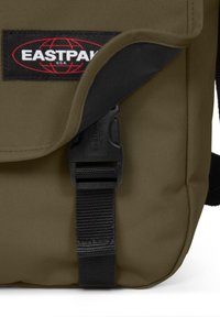 Olivově zelená taška Eastpak s černým popruhem a sponou, s logem na nášivce. Texturovaná tkanina se zavíráním na klopu.