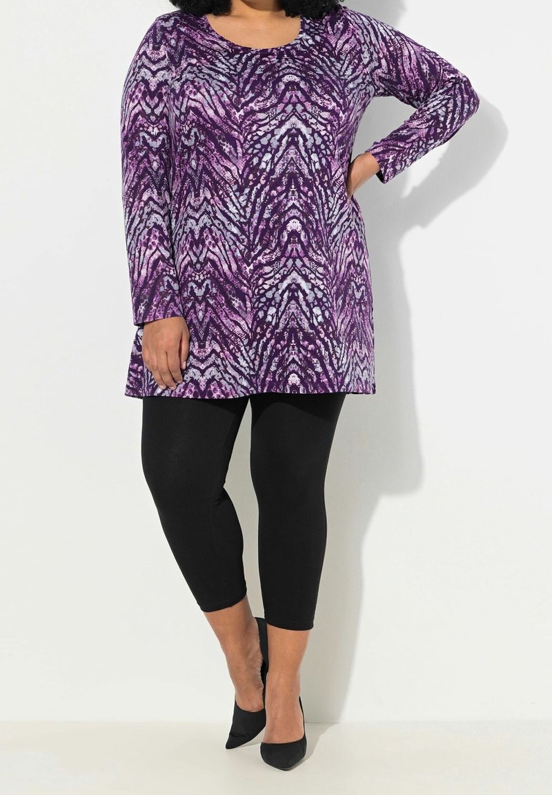 Tunique à manches longues dans un motif abstrait violet et gris, associée à des leggings noirs et des escarpins noirs pointus. Tissu lisse et extensible.