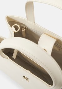 Cremefarbene Ledertasche mit goldenen Ringdetails, offener Oberseite, die ein inneres Reißverschlussfach und einen Riemen mit Magnetverschluss offenbart.