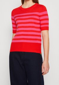 Punainen ja vaaleanpunainen raidallinen lyhythihainen neulepaita, jossa on pyöreä pääntie ja ribbimekka, valmistettu pehmeästä, tekstuurista neulomateriaalista.