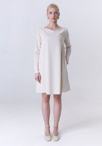 Ein langärmliges, cremefarbenes Kleid mit V-Ausschnitt, geradem Schnitt, seitlichen Taschen und glatter Stoffqualität, kombiniert mit hellen, knöchelhohen Schuhen mit Riemchen.