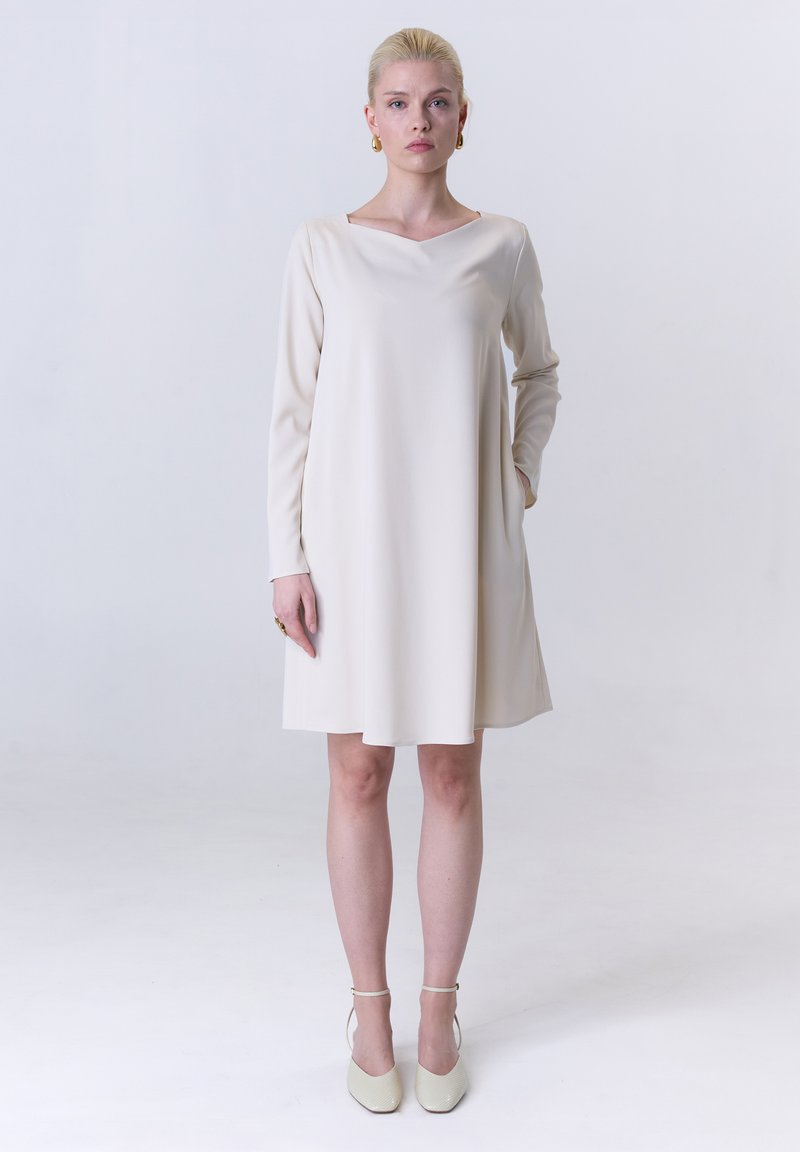 Ein langärmliges, cremefarbenes Kleid mit V-Ausschnitt, geradem Schnitt, seitlichen Taschen und glatter Stoffqualität, kombiniert mit hellen, knöchelhohen Schuhen mit Riemchen.