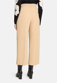 Pantalon beige à jambes larges avec une ceinture côtelée, doté de deux poches arrière. Porté avec des bottes noires montant jusqu'aux genoux. Texture de tissu lisse.