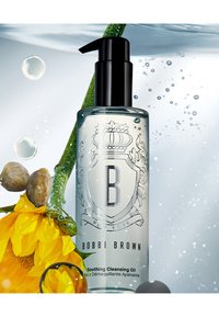 Heldere glazen fles met een zwarte pompkop, gelabeld "Bobbi Brown Soothing Cleansing Oil." Heeft een bloemenontwerp en is omringd door waterdruppels en bloemblaadjes.