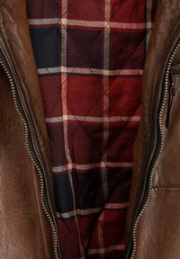 Bruine leren jas met een ritsluiting en een rood en marineblauw geruit gevoerd binnenwerk, met diagonale en horizontale patronen.