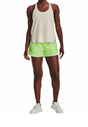 Mujer con top sin mangas beige claro, pantalones cortos deportivos verde neón y zapatillas de correr blancas, de pie contra un fondo blanco.