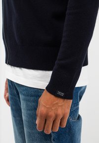 Navy Zip-Up-Strickjacke aus weichem Stoff, mit gerippten Bündchen und Saum, und einem Etikett am Bündchen. Kombiniert mit einem weißen T-Shirt und blauen Jeans.