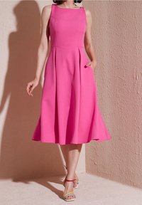 Robe midi rose vif sans manches avec corsage ajusté, jupe évasée et poches latérales portée avec des sandales à brides multicolores.