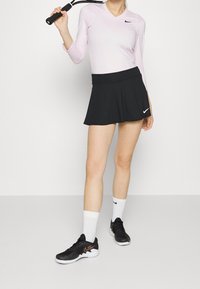 Ljusrosa långärmad topp, svart plisserad kjol, vita strumpor och svarta sportskor med oranget accent. Tennisracket hålls i handen.