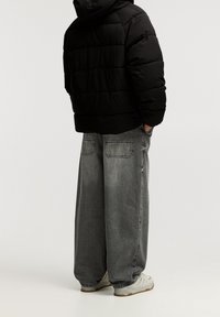 Chaqueta acolchada negra con capucha, combinada con pantalones vaqueros grises de pierna ancha. Detalles destacados incluyen grandes bolsillos traseros y una textura suave. Zapatillas blancas.