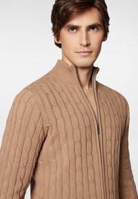 Beżowy sweter robiony na drutach z wysokim kołnierzem, posiadający wzór w warkocze i zamek błyskawiczny. Gładka faktura z prążkowanymi akcentami.