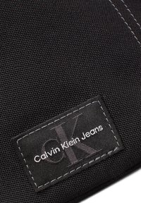 Melna auduma etiķete ar tekstu "Calvin Klein Jeans" un pelēku "CK" logo, ko apņem balta šuvju apmale uz teksturēta fona.