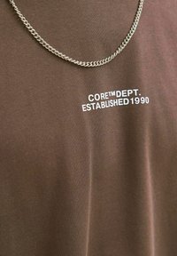 Brun t-shirt med tekstureret stof, der har hvid tekst "CORE™ DEPT. ETABLERT 1990". Akkompagneret af en sølvring halskæde.