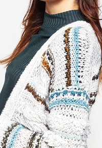 Vit stickad cardigan med flerfärgade texturerade mönster i blått, guld och brunt, lager på lager över en mörkgrön ribbad polotröja.