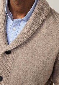 Beżowy kardigan dzianinowy z kołnierzem szalowym, teksturowany materiał i czarne guziki. Noszony na niebieskiej koszuli w paski z klasycznym kołnierzykiem.