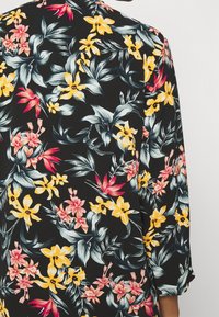 Tissu noir avec des fleurs tropicales jaunes, roses et rouges et des feuilles vertes formant un motif floral vibrant sur un vêtement à manches longues.