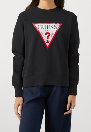 Osoba w czarnej bluzie z czerwonobiałym trójkątnym logo GUESS i granatowych spodniach, stojąca na tle jednolitego tła.