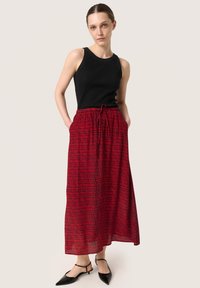 Černý žebrovaný top na ramínka v kombinaci s volnou červenou maxi sukni s texturovaným černým vzorem. Modelka má na sobě černé sandály na podpatku.