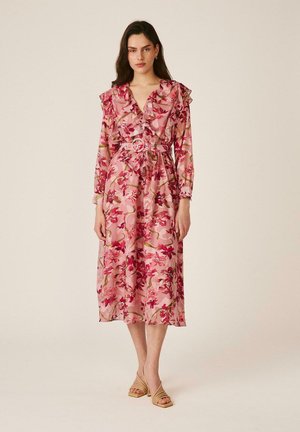 Mujer con vestido midi rosa con flores, volantes y un cinturón a juego, de pie con sandalias beige con tacón sobre un fondo liso.