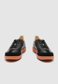 Par sorte lædersneakers med gummisåler i en naturlig farve og sorte snørebånd, set forfra mod en lysegrå baggrund.