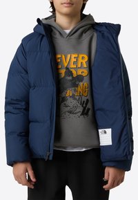 Tumesinine puksjakk üle suuruse, millel on kapuuts, tõmblukk ja vasakul varrukal logoga plekk.