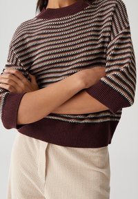 Person with crossed arms wearing a brown, white, and black striped knit sweater and beige corduroy pants.