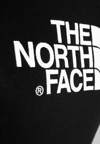 Logo et emblème blancs "THE NORTH FACE" cousus sur un fond en tissu noir texturé.