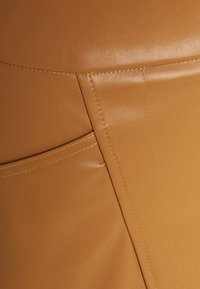 Material de cuero marrón claro con textura suave, que presenta costuras visibles y un detalle de bolsillo lateral, destacando un diseño contorneado y ajustado.