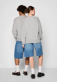 Felpe grigie oversize, pantaloni shorts in denim blu con orli sfilacciati, calze bianche e scarpe scure. Cuciture visibili e dettagli di vestibilità rilassata.