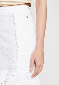 Bílá sukně vyrobená z texturované látky s výšivkovými vzory, volánovými okraji a stříbrným kovovým detailem na pase.