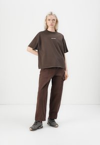 Barna pamut, oversized póló fehér logóval, barna nadrággal és szürke sportcipővel párosítva, egy sima fehér háttér előtt áll.