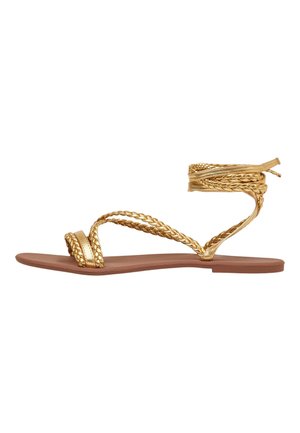 Flache Sandale mit geflochtenen metallic-goldenen Riemen, Zehensteg und Wickelriemen um den Knöchel auf einer braunen Sohle, seitlich betrachtet.